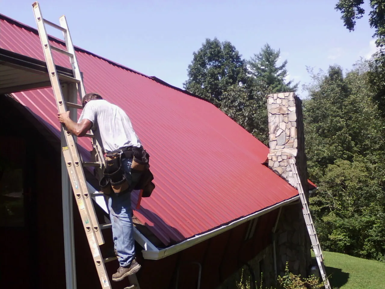 Roofer on a ladder beside a red metal roof and stone chimney — Soffit & Fascia Repair by Roofing Brothers in Schaumburg